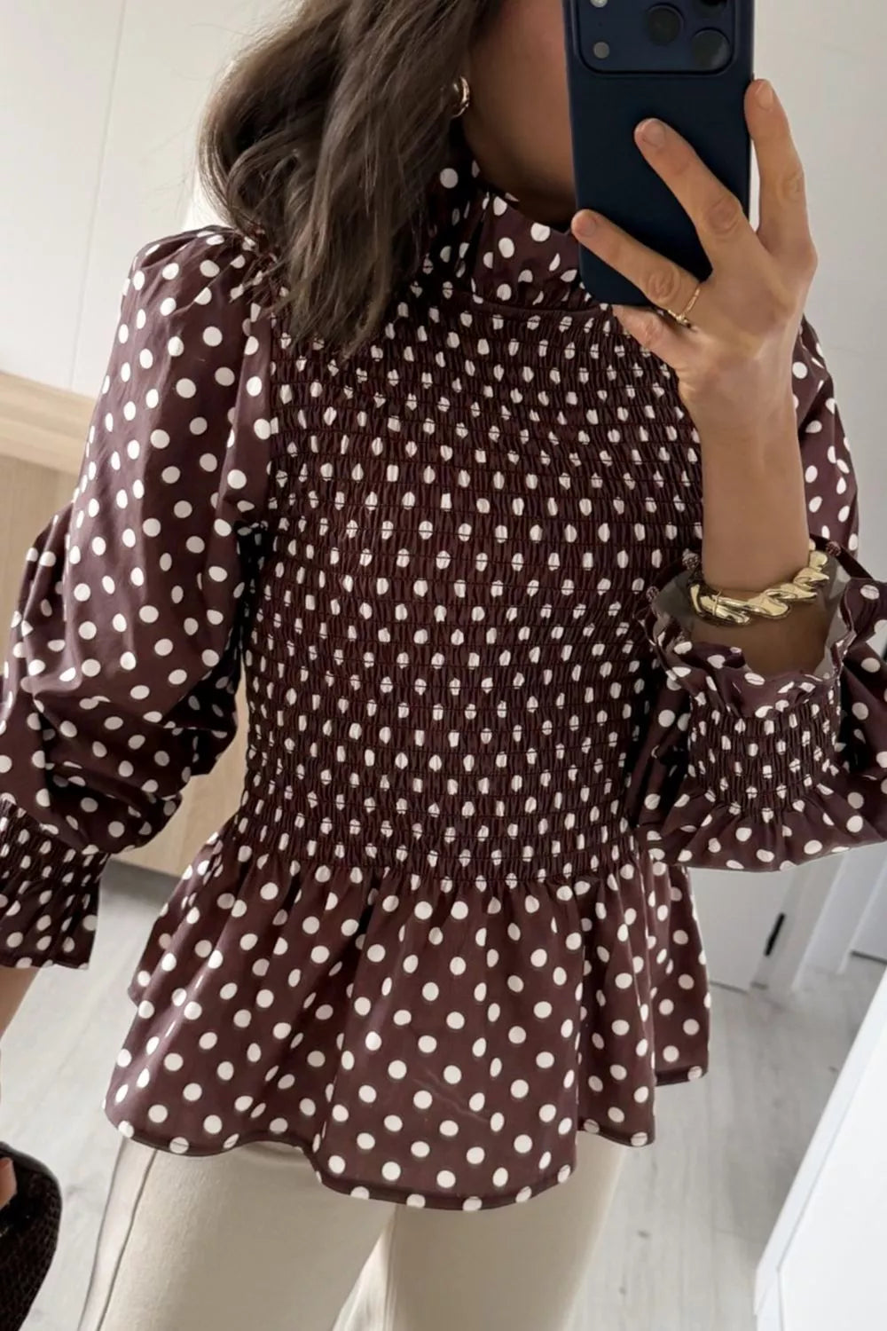 Vista frontal de blusa marrón con lunares blancos, cuello alto, cintura fruncida y mangas abullonadas con puños de volantes. NINA AURA COLLECTION.